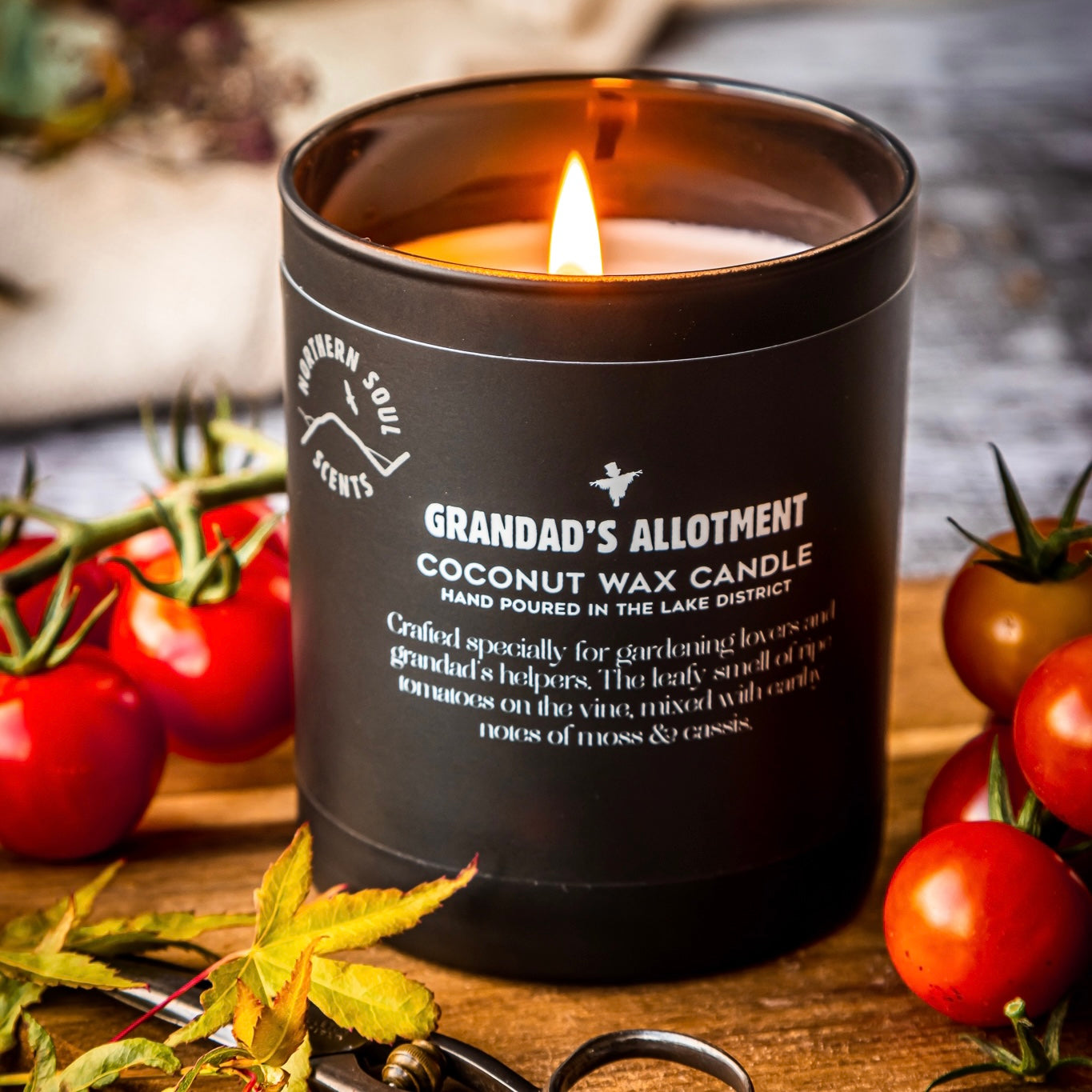 Grandad's Allotment scented candle in a matte black opaque candle jar with a scarecrow and tomatoe design on premium quality labelling for a sleek look. Perfect for gifting.